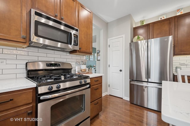 kitchen with brown cabinetry, backsplash, dark wood-style flooring, light stone counters, and stainless steel appliances