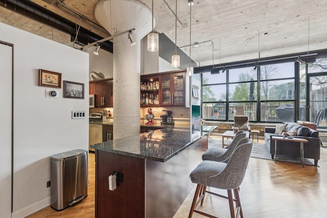 kitchen featuring track lighting, light wood finished floors, dark stone countertops, a kitchen bar, and glass insert cabinets