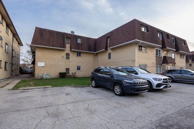 exterior space with brick siding, a shingled roof, uncovered parking, and mansard roof