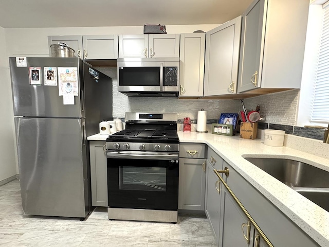 kitchen featuring light stone countertops, a sink, stainless steel appliances, decorative backsplash, and gray cabinetry