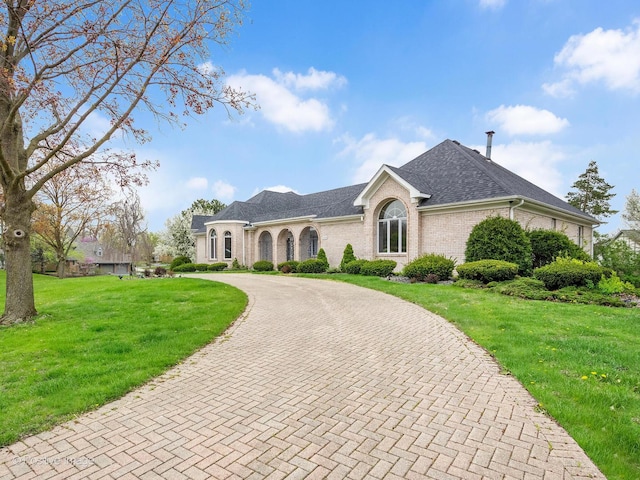 french country inspired facade featuring curved driveway, a shingled roof, a front yard, and brick siding