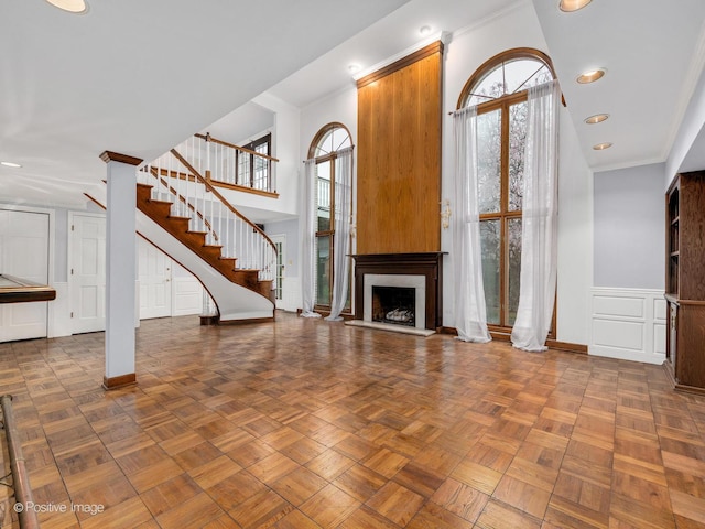unfurnished living room featuring wainscoting, a decorative wall, a fireplace with raised hearth, stairs, and ornamental molding