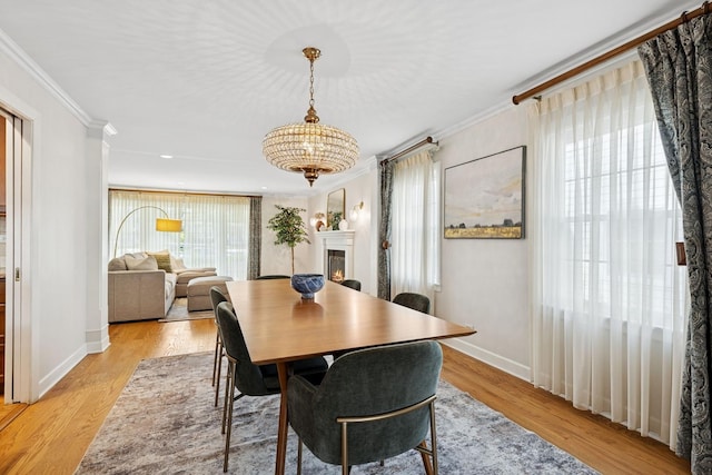 dining space with a warm lit fireplace, a chandelier, baseboards, light wood-type flooring, and ornamental molding