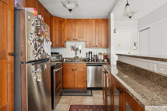 kitchen with a sink, stainless steel appliances, light tile patterned floors, dark stone countertops, and brown cabinetry