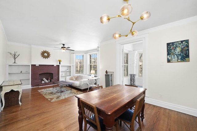 dining area with wood finished floors, ceiling fan with notable chandelier, a brick fireplace, baseboards, and crown molding