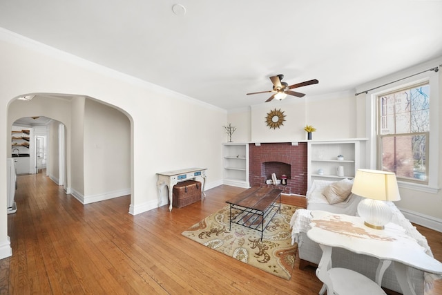 living room with a brick fireplace, ceiling fan, baseboards, arched walkways, and hardwood / wood-style flooring