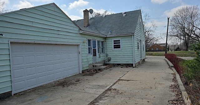 rear view of property with concrete driveway, a shingled roof, an attached garage, and a chimney