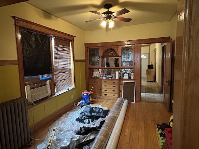 bedroom featuring wood finished floors, radiator heating unit, cooling unit, a wainscoted wall, and a ceiling fan