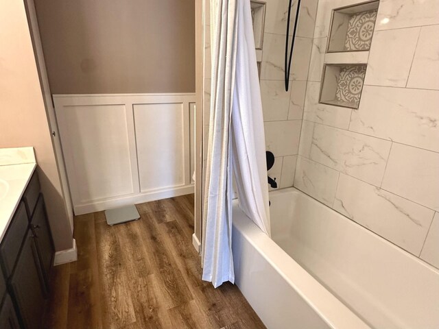 bathroom featuring wainscoting, a decorative wall, vanity, shower / bathtub combination with curtain, and wood finished floors