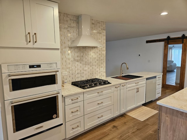 kitchen featuring wood finished floors, white double oven, gas cooktop, a sink, and wall chimney exhaust hood