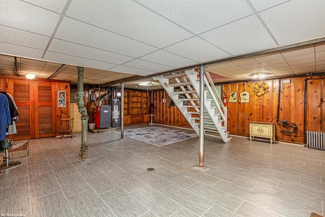finished below grade area featuring wooden walls, gas water heater, radiator heating unit, a drop ceiling, and stairway