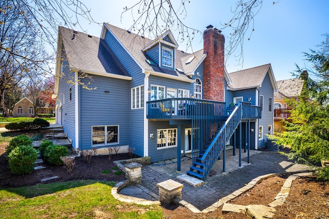 rear view of property with a deck, roof with shingles, a chimney, a patio area, and stairway
