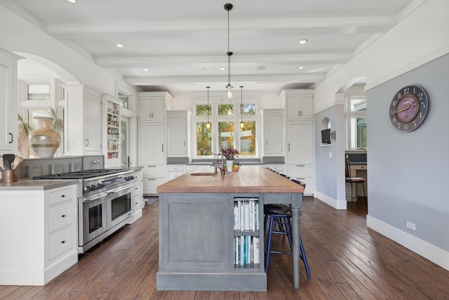 kitchen featuring white cabinets, beam ceiling, a kitchen island with sink, arched walkways, and range with two ovens