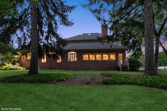 back of property at dusk with a shingled roof, a chimney, brick siding, and a lawn