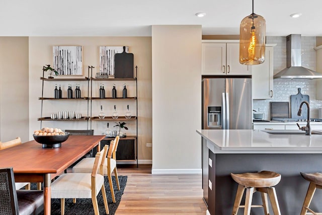 kitchen featuring wall chimney range hood, stainless steel fridge, light wood-style flooring, white cabinetry, and backsplash