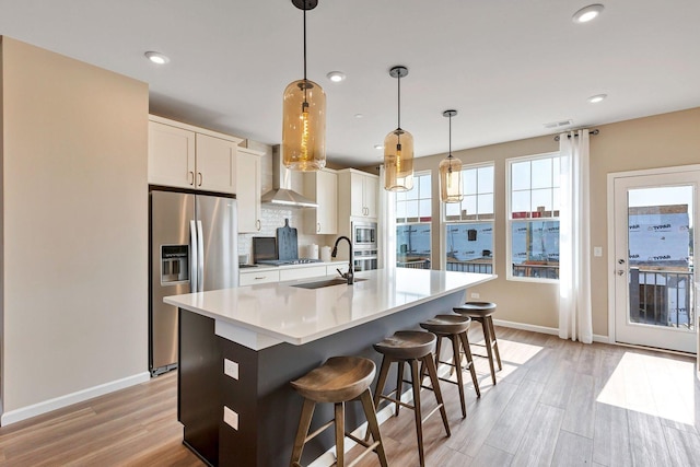 kitchen featuring a kitchen island with sink, appliances with stainless steel finishes, a sink, baseboards, and wall chimney exhaust hood