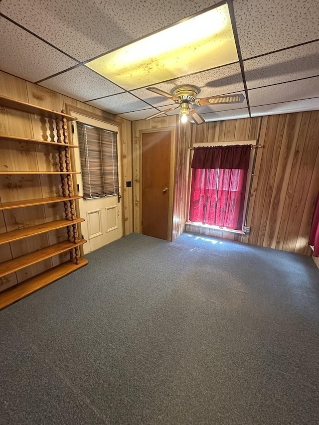 carpeted empty room with a paneled ceiling, ceiling fan, and wooden walls