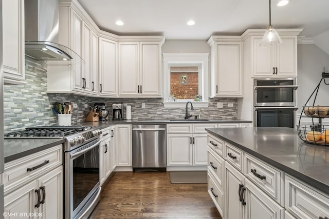kitchen with dark countertops, appliances with stainless steel finishes, a sink, wall chimney exhaust hood, and tasteful backsplash