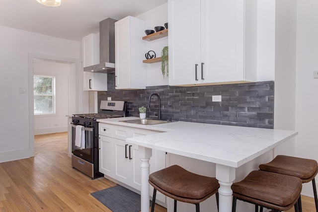 kitchen featuring wall chimney range hood, stainless steel gas range oven, open shelves, a sink, and a breakfast bar