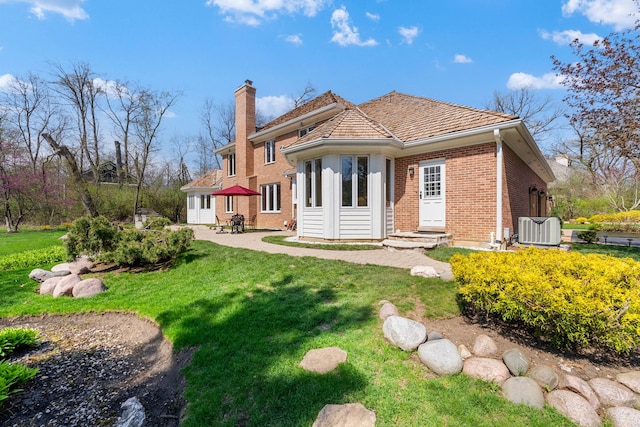 rear view of property featuring brick siding, a chimney, a yard, cooling unit, and a patio