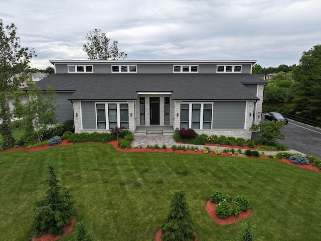 view of front facade with roof with shingles, a front yard, and stone siding