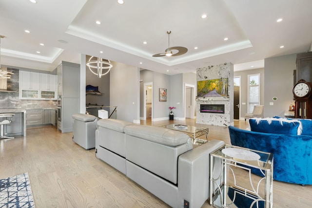 living room with a raised ceiling, a large fireplace, light wood-style flooring, a ceiling fan, and recessed lighting