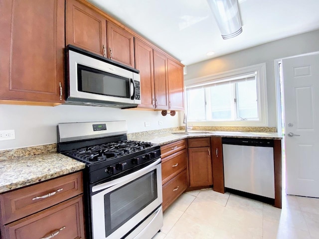 kitchen featuring brown cabinetry, appliances with stainless steel finishes, a sink, light tile patterned floors, and light stone counters