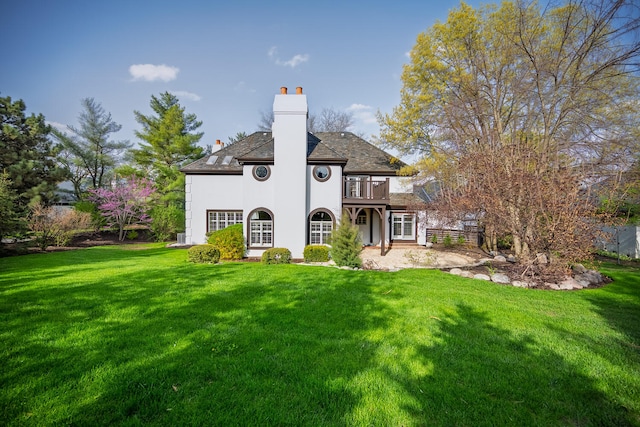 back of house with a lawn, stucco siding, a balcony, and a chimney