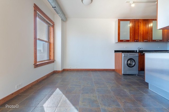 kitchen with dark countertops, a sink, brown cabinetry, baseboards, and washer / dryer