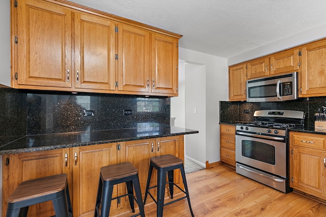 kitchen with decorative backsplash, a breakfast bar area, appliances with stainless steel finishes, light wood-type flooring, and dark stone counters