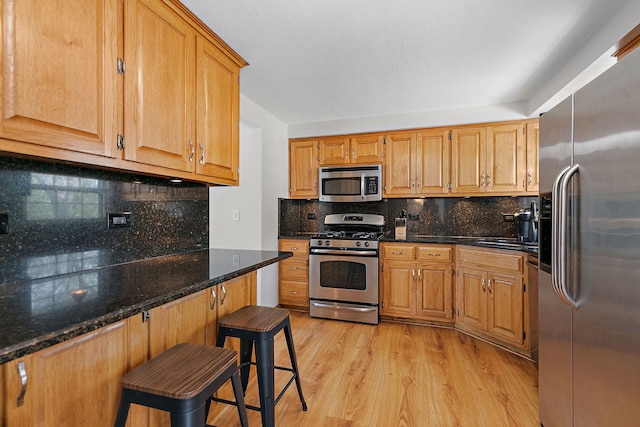 kitchen with light wood finished floors, a breakfast bar area, dark stone counters, stainless steel appliances, and tasteful backsplash
