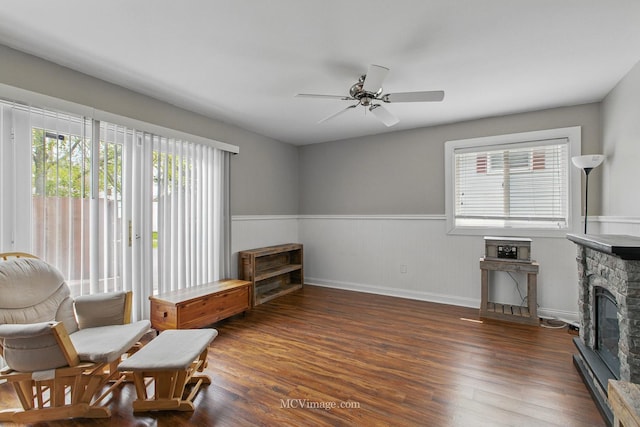 living area featuring wainscoting, ceiling fan, a stone fireplace, and wood finished floors