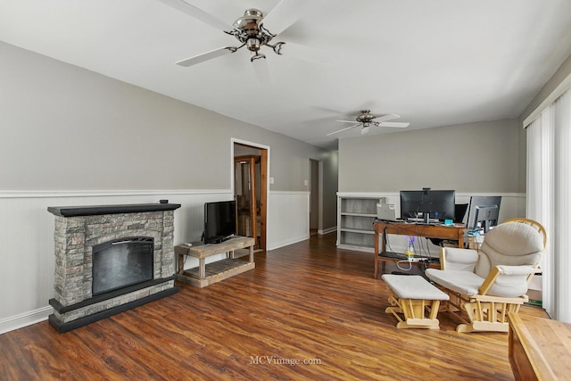 living area featuring a ceiling fan, a stone fireplace, wainscoting, and wood finished floors