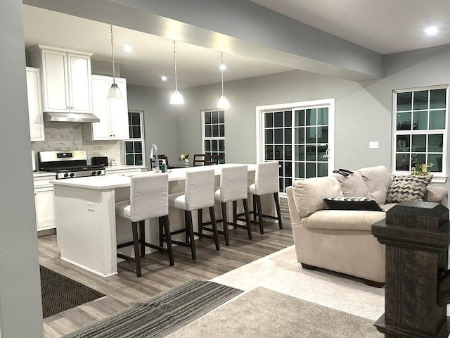 kitchen with a kitchen island with sink, backsplash, stainless steel range with gas stovetop, wood finished floors, and under cabinet range hood