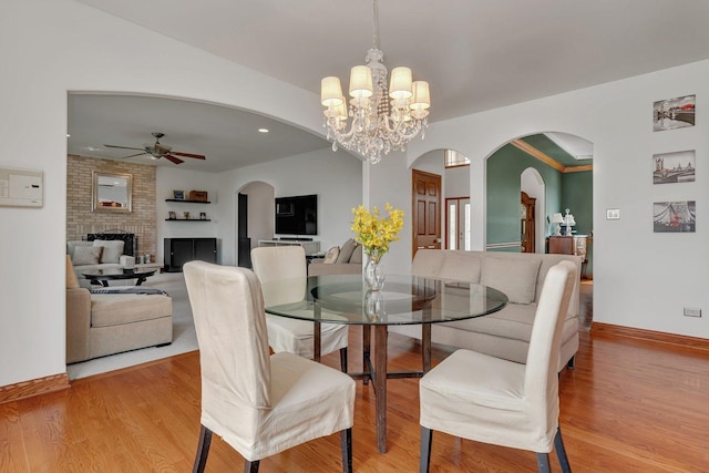 dining space with light wood-style floors, arched walkways, a brick fireplace, and ceiling fan with notable chandelier