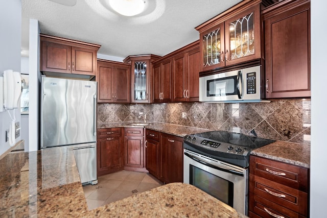 kitchen with appliances with stainless steel finishes, a sink, dark stone counters, tasteful backsplash, and glass insert cabinets