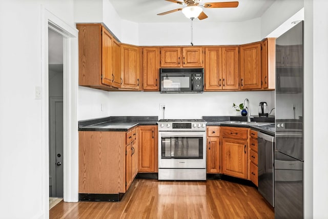 kitchen featuring brown cabinetry, a ceiling fan, dark countertops, appliances with stainless steel finishes, and wood finished floors