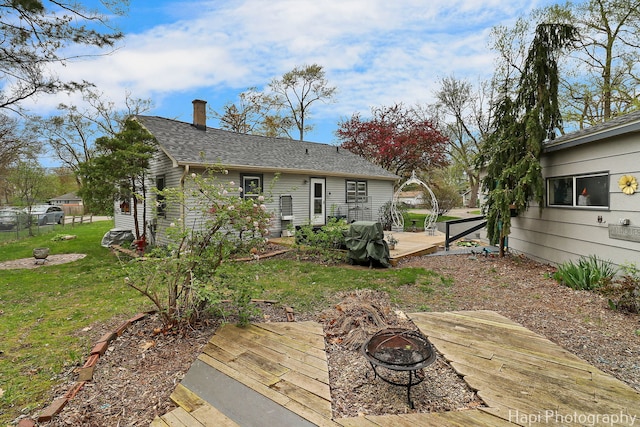 back of house featuring a chimney, a lawn, a deck, a shingled roof, and a fire pit