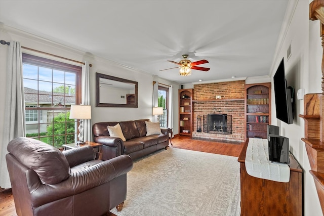 living room with a ceiling fan, visible vents, a brick fireplace, wood finished floors, and ornamental molding