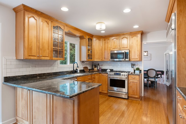 kitchen featuring appliances with stainless steel finishes, a sink, dark stone counters, a peninsula, and glass insert cabinets