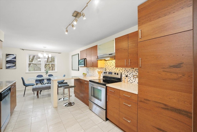 kitchen with under cabinet range hood, brown cabinets, a chandelier, tasteful backsplash, and appliances with stainless steel finishes