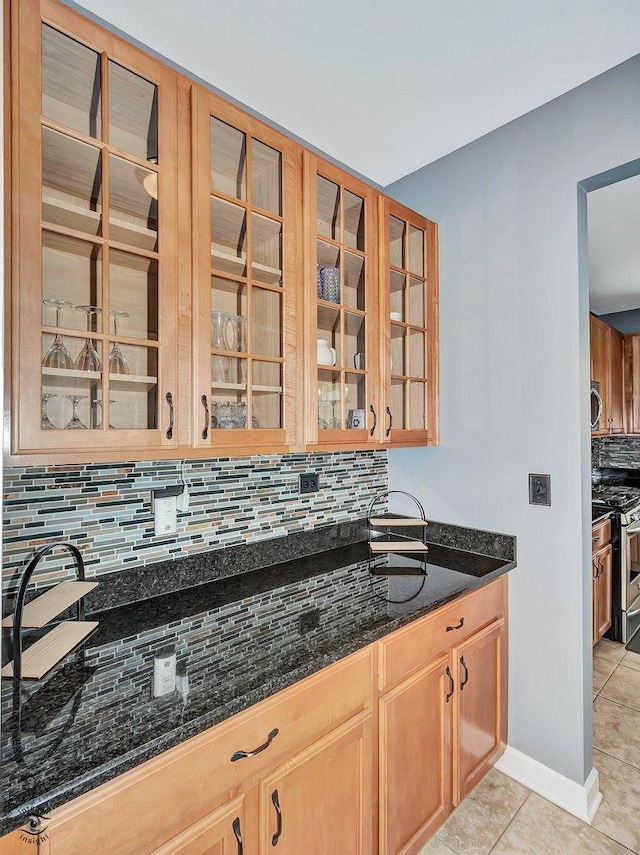 kitchen featuring stainless steel gas stove, dark stone counters, light tile patterned floors, tasteful backsplash, and glass insert cabinets