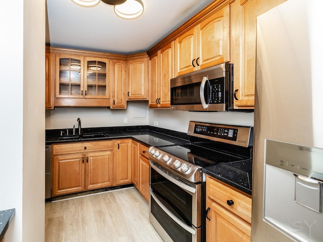 kitchen with appliances with stainless steel finishes, a sink, glass insert cabinets, dark stone counters, and light wood-style flooring