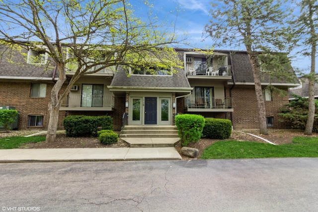 view of property with brick siding, roof with shingles, a balcony, and mansard roof