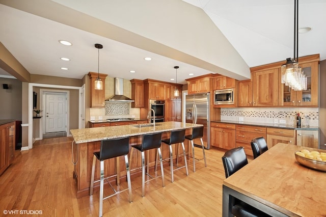 kitchen with brown cabinetry, wall chimney range hood, a kitchen bar, light wood finished floors, and stainless steel appliances