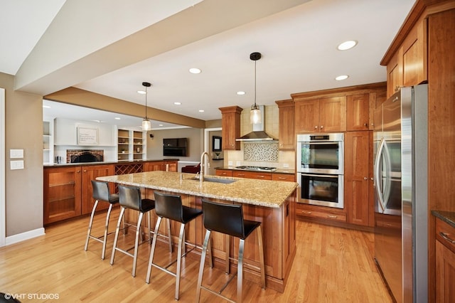kitchen with appliances with stainless steel finishes, brown cabinets, a sink, open floor plan, and wall chimney exhaust hood
