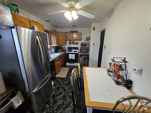 kitchen featuring ceiling fan, tile countertops, brown cabinets, a sink, and appliances with stainless steel finishes
