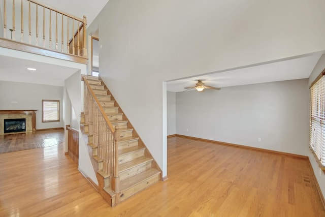 staircase with a ceiling fan, wood finished floors, baseboards, a glass covered fireplace, and a high ceiling