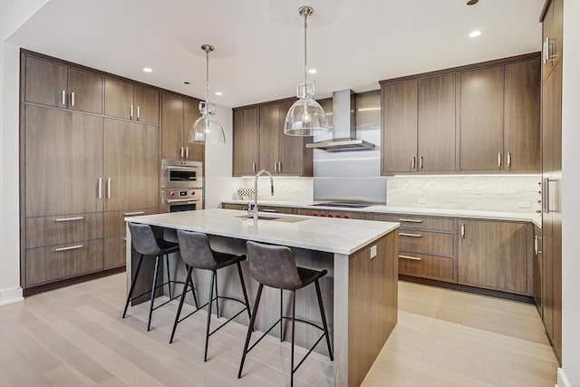 kitchen featuring wall chimney exhaust hood, a center island with sink, a sink, a breakfast bar, and light wood-style flooring