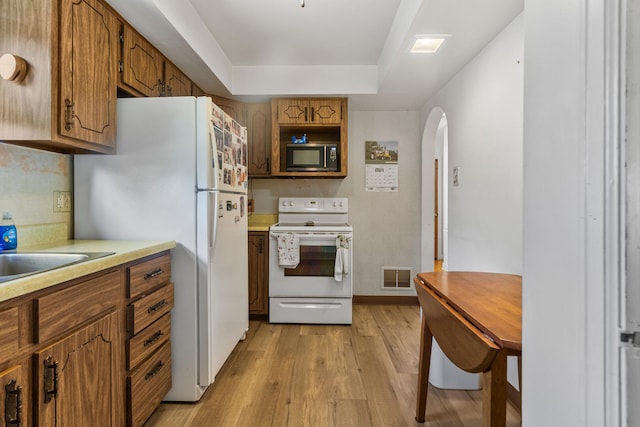 kitchen featuring white appliances, arched walkways, light countertops, light wood finished floors, and brown cabinetry